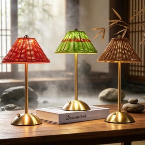 May include: Three decorative table lamps with woven shades in red, green, and brown. Each lamp has a gold-colored base and stem. The lamps are arranged on a wooden surface, with a book titled "Discontent" visible in the center.