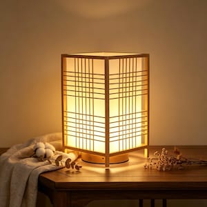 May include: A rectangular wooden table lamp with a grid-like design. The lamp has a warm, soft glow, and the frame is made of light-colored wood. The lamp is on a wooden table with a neutral-colored cloth and dried flowers.