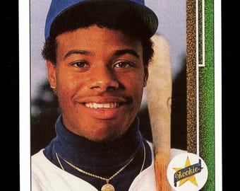 May include: A 1989 Upper Deck baseball card featuring Ken Griffey Jr. wearing a blue cap with a yellow S, a dark blue turtleneck, and a gold chain. He holds a wooden baseball bat. The card includes a yellow star Rookie logo and his name.