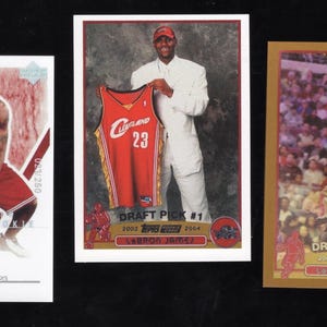 May include: Three rectangular sports trading cards featuring LeBron James. The left card shows him in a red Cavaliers uniform with a signature. The center card displays him holding a jersey. The right card depicts him shooting a basketball.
