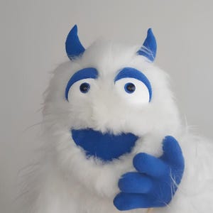 May include: A white plush monster puppet with long faux fur, featuring bright blue horns, blue eyebrows, blue eyes, and a wide blue mouth. The puppet has a blue hand raised in a waving gesture and is attached to a thin wooden stick for manipulation.