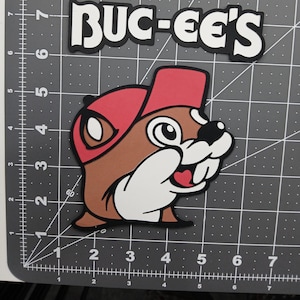Puede incluir: Una colorida pegatina con el logotipo de Buc-ee's que presenta un castor de dibujos animados con una gorra roja. El castor tiene un cuerpo marrón, dientes blancos y una expresión sonriente. La pegatina también incluye el texto "BUC-EE'S" en letras blancas con un contorno negro.