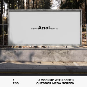 Op de afbeelding: Een grote, lege outdoor mega screen mockup met de tekst "Studio Arial Mockup" in het zwart. Het scherm is omlijst in metaal en geplaatst tegen een betonnen muur en een bosachtergrond. De onderkant van de afbeelding bevat de tekst "1 PSD" en "<MOCKUP WITH SCNE> OUTDOOR MEGA SCREEN".