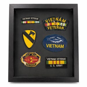May include: A black shadow box frame containing six embroidered military patches and ribbons. The collection includes Vietnam Veteran insignia, a 1st Cavalry Division patch, a helicopter graphic, and a U.S. Army Vietnam Veteran service patch.