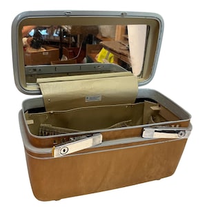 May include: A tan Samsonite Profile hard-shell train case with a silver-toned frame and dual chrome-finished latches. The lid features an interior mirror, and the main compartment is lined with tan fabric, including a small elasticized pocket.