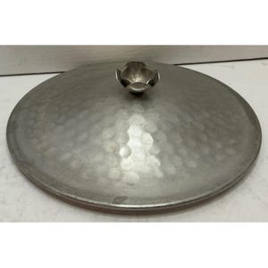 May include: A circular, silver-toned metal lid featuring a hammered texture across its surface. The lid is topped with a small, three-dimensional metal flower-shaped handle centered at the top, providing a decorative grip for the flat cover.