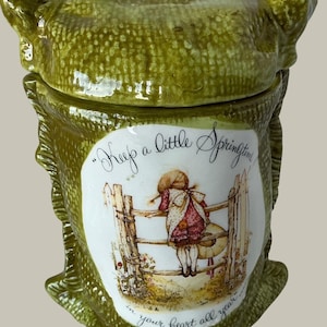 May include: A green ceramic jar with a textured surface and a lid. The front features a white oval illustration of a person leaning on a wooden fence, with the text "Keep a little Springtime in your heart all year".
