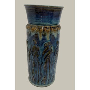 May include: A cylindrical ceramic vase featuring a flared rim and a deep blue glaze with brown drip accents. The body displays a textured, vertical relief pattern with overlapping blue and brown tones, creating a layered, glossy finish.