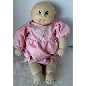 May include: A vintage doll with a bald head, blue eyes, and a puckered expression. The doll is wearing a pink dress with puffy sleeves and teal trim. The doll's arms and legs are made of a soft, beige material.