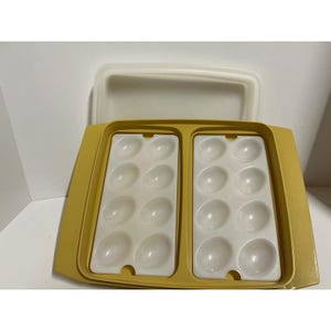 May include: A rectangular yellow plastic egg storage container with a removable lid. The base features two white plastic inserts, each containing eight individual oval-shaped indentations designed to hold sixteen eggs in total.