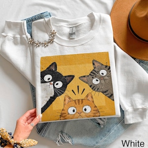 Puede incluir: Una sudadera blanca de cuello redondo con un estampado gráfico de tres gatos con ojos grandes sobre un fondo amarillo. Los gatos incluyen un gato blanco y negro, un gato atigrado gris y un gato atigrado naranja. La sudadera tiene mangas largas.