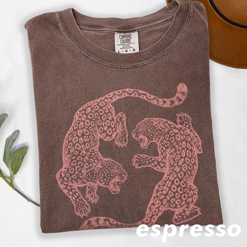 May include: Espresso-colored t-shirt featuring a pink leopard design. The shirt has a crew neck and short sleeves. The graphic shows two leopards facing each other. The shirt is folded and has a tag that says "Comfort Colors".