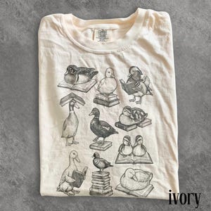 Puede incluir: Camiseta de color marfil con un gr&aacute;fico de patos leyendo libros. Los patos est&aacute;n en varias poses, algunos sentados, otros de pie y algunos leyendo. La camiseta tiene el logo "Comfort Colors" en la parte superior izquierda y la palabra "ivory" en la parte inferior derecha.