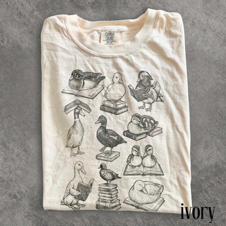 Puede incluir: Camiseta de color marfil con un gr&aacute;fico de patos leyendo libros. Los patos est&aacute;n en varias poses, algunos sentados, otros de pie y algunos leyendo. La camiseta tiene el logo "Comfort Colors" en la parte superior izquierda y la palabra "ivory" en la parte inferior derecha.