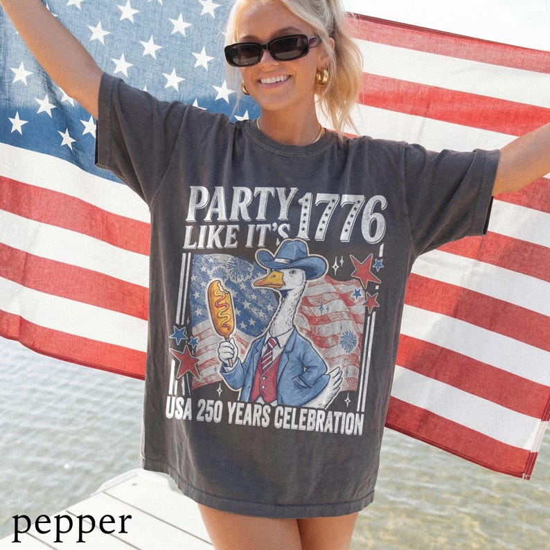 May include: Dark gray t-shirt with the text "PARTY LIKE IT'S 1776" and a graphic of a goose in a cowboy hat holding a corn dog. The shirt celebrates the USA's 250th anniversary. An American flag is in the background.