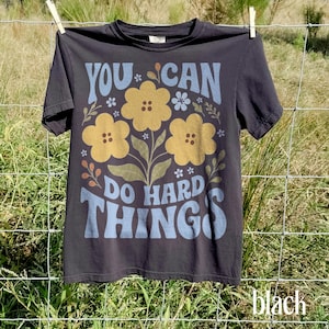 May include: A black short-sleeve t-shirt featuring a graphic print with the text YOU CAN DO HARD THINGS in light blue. The design includes three large yellow flowers with brown centers, small light blue flowers, and green leaves and stems.