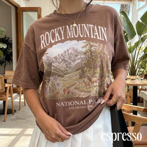 Puede incluir: Camiseta marrón con un gráfico del Parque Nacional de las Montañas Rocosas en Colorado, EE. UU. El diseño presenta una cordillera, árboles, un río y ciervos. El texto "ROCKY MOUNTAIN" y "NATIONAL PARK" está impreso.