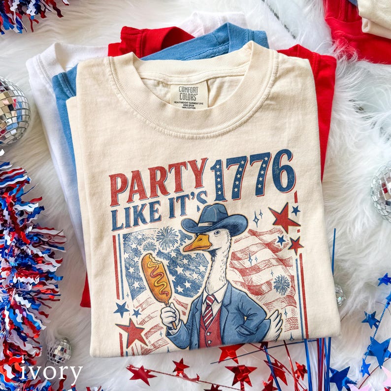 May include: Ivory-colored t-shirt with the text "PARTY 1776 LIKE IT'S" and a cartoon goose wearing a cowboy hat and suit, holding a corn dog. The shirt is stacked with red, white, and blue shirts.