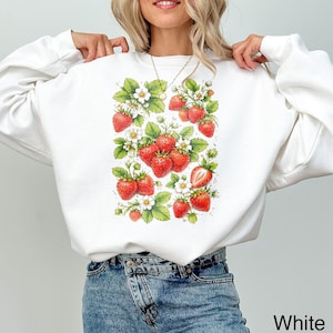 May include: A white long-sleeve sweatshirt featuring a central graphic print of multiple red strawberries, green leaves, and small white flowers. The design is arranged in a rectangular cluster on the front of the garment, showcasing various ripe strawberries in different sizes and orientations.