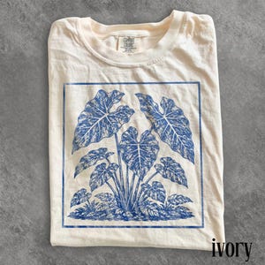 May include: An ivory t-shirt featuring a blue graphic print of a cluster of large, heart-shaped leaves within a rectangular border. The shirt has a crew neckline and a Comfort Colors brand label printed on the inside of the collar area.