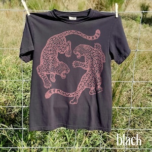 May include: Black t-shirt featuring a pink leopard graphic. The design shows two leopards in a dynamic pose. The shirt is hanging on a clothesline. The word "black" is printed at the bottom of the image.