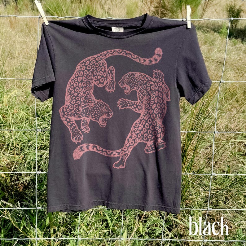 May include: Black t-shirt featuring a pink leopard graphic. The design shows two leopards in a dynamic pose. The shirt is hanging on a clothesline. The word "black" is printed at the bottom of the image.