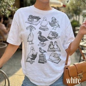 Puede incluir: Camiseta blanca con ilustraciones de patos y libros. Los patos est&aacute;n representados en diferentes poses, algunos leyendo, otros descansando sobre libros. La palabra "white" est&aacute; impresa en la esquina inferior derecha.