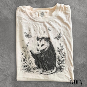 May include: An ivory t-shirt featuring a black ink illustration of an opossum reading a book, surrounded by flowers, butterflies, stars, and a crescent moon. The shirt has a crew neckline and a Comfort Colors brand label at the inner collar.