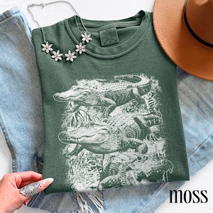 Op de afbeelding: Een mosgroen t-shirt met een witte afbeelding van alligators. Het shirt heeft een ronde hals en korte mouwen. Het woord "moss" staat rechtsonder in het wit gedrukt. Een zilveren ketting en een bruine hoed zijn ook zichtbaar.