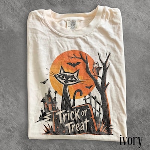May include: An ivory t-shirt featuring a graphic print of a black cat, a large orange moon, a haunted house, an owl in a tree, and bats. The text Trick or Treat is printed in black lettering at the bottom of the design, with a Comfort Colors label.