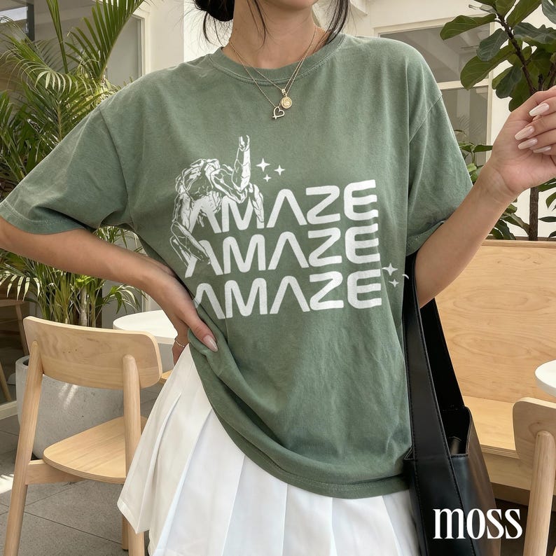 May include: Olive green t-shirt with the word "AMAZE" stacked three times in white, along with a graphic and stars. The shirt is paired with a white pleated skirt and a black tote bag. The word "moss" is in the bottom right corner.