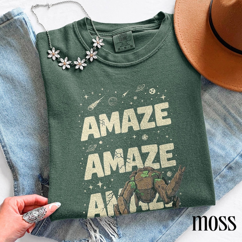May include: A moss green t-shirt with the word "AMAZE" repeated three times in a cream-colored font, along with a graphic of a space-themed character. A silver necklace with flower-shaped pendants and a brown hat are also visible.