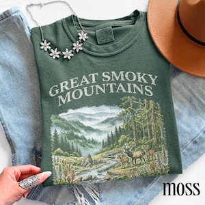 May include: Moss green t-shirt with the text "GREAT SMOKY MOUNTAINS" and a scenic mountain and forest graphic. The shirt is paired with blue jeans, a silver necklace, and a brown hat.