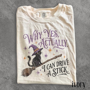 May include: An ivory t-shirt featuring a graphic of a black cat wearing a purple witch hat, sitting on a broomstick. The text reads, "WHY YES, ACTUALLY, I CAN DRIVE A STICK." Small purple and orange stars surround the cat and the text.
