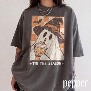 May include: A charcoal grey short-sleeve t-shirt featuring a graphic of a ghost wearing a witch hat and holding a pumpkin-themed drink. The background of the graphic is a checkered orange and white pattern with bats. The text reads TIS THE SEASON.
