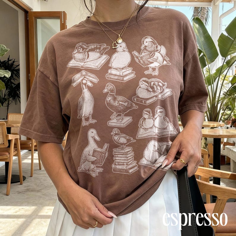 Puede incluir: Camiseta marr&oacute;n con un estampado repetido de patos sobre libros. Los patos est&aacute;n representados en diferentes poses, algunos leyendo, otros descansando. La palabra "espresso" est&aacute; impresa en blanco en la esquina inferior derecha de la imagen.