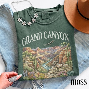 May include: A moss green t-shirt with a Grand Canyon graphic. The design features a detailed landscape with a river, cacti, and wildlife. A silver flower necklace and a brown hat are also visible.