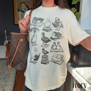 Puede incluir: Camiseta de color marfil con un patr&oacute;n repetido de patos sobre libros. Los patos est&aacute;n representados en varias poses, algunos leyendo, otros descansando. La palabra "ivory" est&aacute; impresa en peque&ntilde;o en la parte inferior derecha.