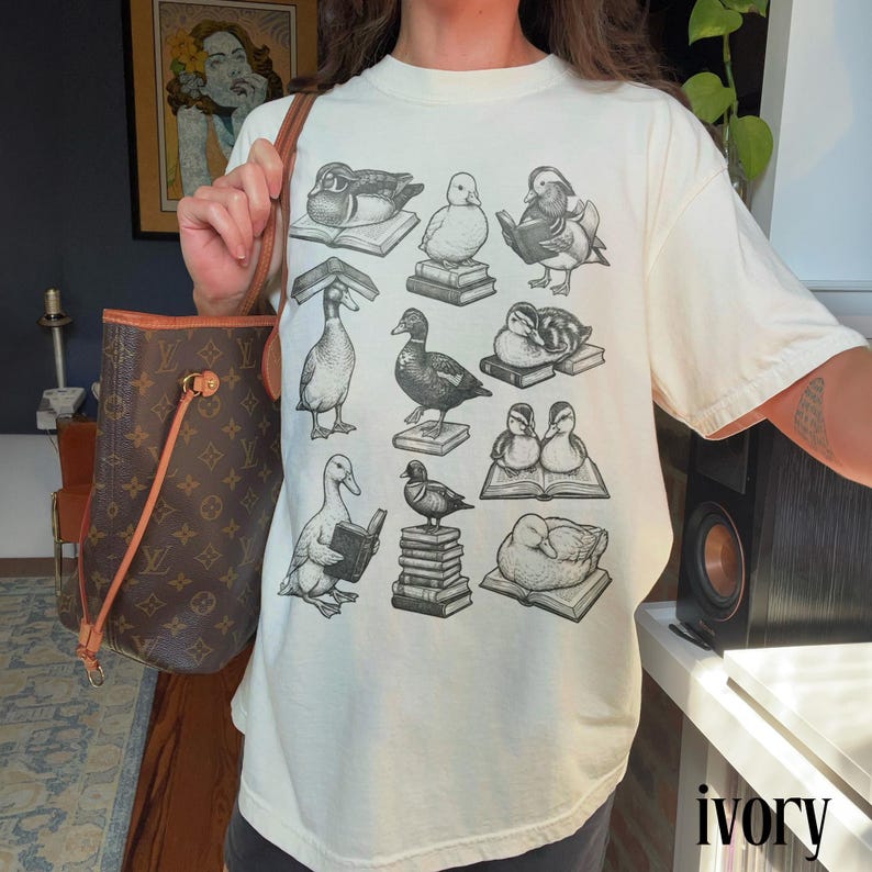 Puede incluir: Camiseta de color marfil con un patr&oacute;n repetido de patos sobre libros. Los patos est&aacute;n representados en varias poses, algunos leyendo, otros descansando. La palabra "ivory" est&aacute; impresa en peque&ntilde;o en la parte inferior derecha.