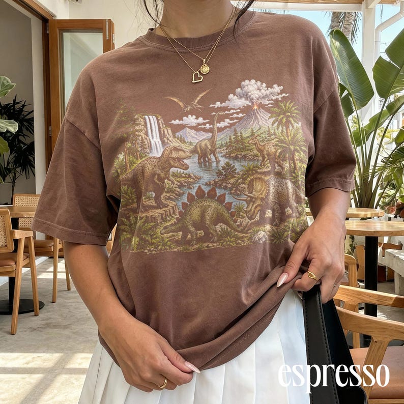 Op de afbeelding: Bruin t-shirt met een gedetailleerd prehistorisch landschap met dinosaurussen, een waterval en een vulkaan. Het shirt heeft korte mouwen en een relaxte pasvorm. Het woord "espresso" staat rechtsonder gedrukt.