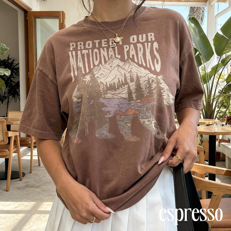 May include: Espresso-colored t-shirt with the text "PROTECT OUR NATIONAL PARKS" above a graphic of a bear, mountains, and trees. The shirt has short sleeves and a relaxed fit.