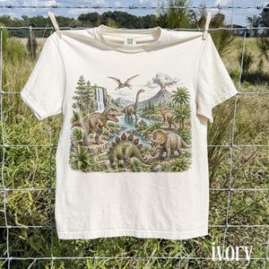 Op de afbeelding: Ivoor kleurig t-shirt met een gedetailleerde illustratie van dinosaurussen in een prehistorisch landschap. Het ontwerp bevat een Tyrannosaurus Rex, Stegosaurus en Triceratops bij een rivier, met een vulkaan en ptero-dactylen op de achtergrond. Het shirt hangt aan een waslijn.