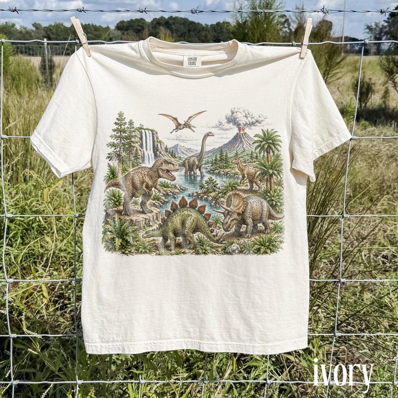 Op de afbeelding: Ivoor kleurig t-shirt met een gedetailleerde illustratie van dinosaurussen in een prehistorisch landschap. Het ontwerp bevat een Tyrannosaurus Rex, Stegosaurus en Triceratops bij een rivier, met een vulkaan en ptero-dactylen op de achtergrond. Het shirt hangt aan een waslijn.