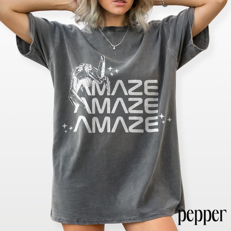 May include: Dark gray t-shirt with the word "AMAZE" repeated three times in white, along with a graphic and star accents. The shirt has short sleeves and a relaxed fit, suitable for casual wear.
