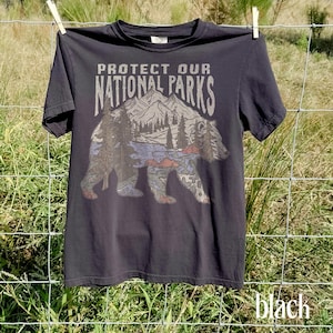 May include: Black t-shirt with the text "PROTECT OUR NATIONAL PARKS" above a graphic of a bear with a mountain and forest scene. The shirt is hanging on a clothesline. The word "black" is written in the bottom right corner.