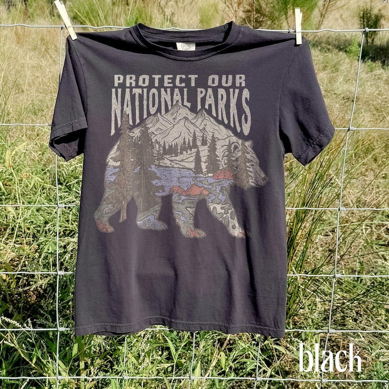 May include: Black t-shirt with the text "PROTECT OUR NATIONAL PARKS" above a graphic of a bear with a mountain and forest scene. The shirt is hanging on a clothesline. The word "black" is written in the bottom right corner.