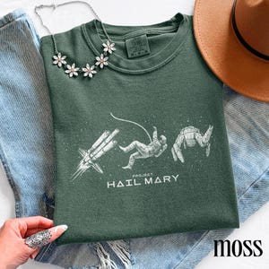 May include: Dark green crew neck sweatshirt featuring a white graphic of a rocket, astronaut, and space crab. The text "PROJECT HAIL MARY" is printed below the graphic. A floral necklace and a brown hat are also visible.