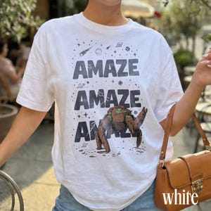 May include: White t-shirt with the word "AMAZE" printed three times in dark gray, along with a space-themed graphic. The bottom graphic is a brown and green creature. The word "white" is printed at the bottom of the image.