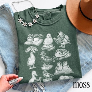 Puede incluir: Una camiseta verde musgo con un gr&aacute;fico blanco de patos sobre libros. La camiseta tiene cuello redondo y mangas cortas. El texto "Comfort Colors" es visible en la etiqueta. Una cadena plateada con detalles florales y un sombrero marr&oacute;n tambi&eacute;n est&aacute;n en la imagen.