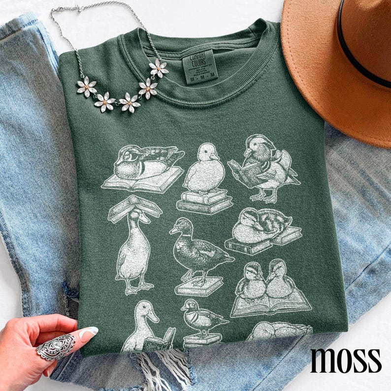 Puede incluir: Una camiseta verde musgo con un gr&aacute;fico blanco de patos sobre libros. La camiseta tiene cuello redondo y mangas cortas. El texto "Comfort Colors" es visible en la etiqueta. Una cadena plateada con detalles florales y un sombrero marr&oacute;n tambi&eacute;n est&aacute;n en la imagen.